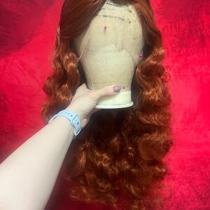 May include: A long, wavy, copper-red wig styled with a side part and voluminous curls. The wig is secured onto a beige mannequin head with several colourful sewing pins inserted into the lace front hairline to hold the hair in place.