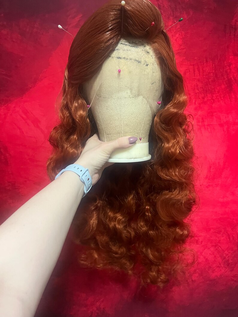 May include: A long, wavy, copper-red wig styled with a side part and voluminous curls. The wig is secured onto a beige mannequin head with several colourful sewing pins inserted into the lace front hairline to hold the hair in place.