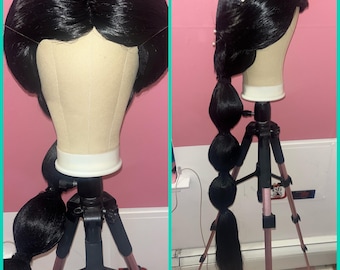 Può includere: Una parrucca nera con una lunga coda di cavallo in stile con più grandi e rotondi boccoli di capelli. La parrucca è su una testa di manichino montata su un treppiede.