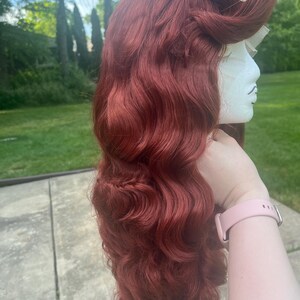 May include: A long, wavy auburn wig styled with a voluminous pompadour at the crown and secured with two small white pearl-tipped pins. The hair features deep, flowing waves that cascade down the side, displayed on a white mannequin head form.