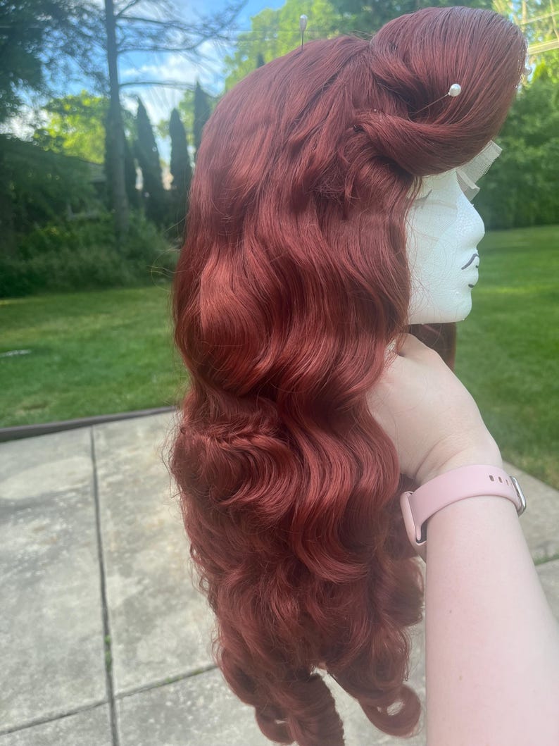 May include: A long, wavy auburn wig styled with a voluminous pompadour at the crown and secured with two small white pearl-tipped pins. The hair features deep, flowing waves that cascade down the side, displayed on a white mannequin head form.