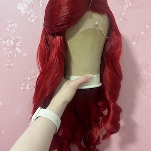 May include: A vibrant red wig with long, wavy hair styled with a side-swept fringe. The wig is secured on a beige mannequin head with several white-tipped pins inserted along the hairline and crown, showcasing a lace front construction.