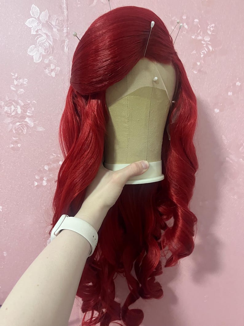 May include: A vibrant red wig with long, wavy hair styled with a side-swept fringe. The wig is secured on a beige mannequin head with several white-tipped pins inserted along the hairline and crown, showcasing a lace front construction.
