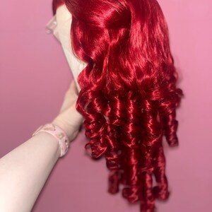 May include: A vibrant, deep red wig featuring long, voluminous hair styled with tight, defined ringlet curls. The hair is parted on the side, showcasing a lace front construction at the hairline, with the curls cascading down in a layered, textured fashion.