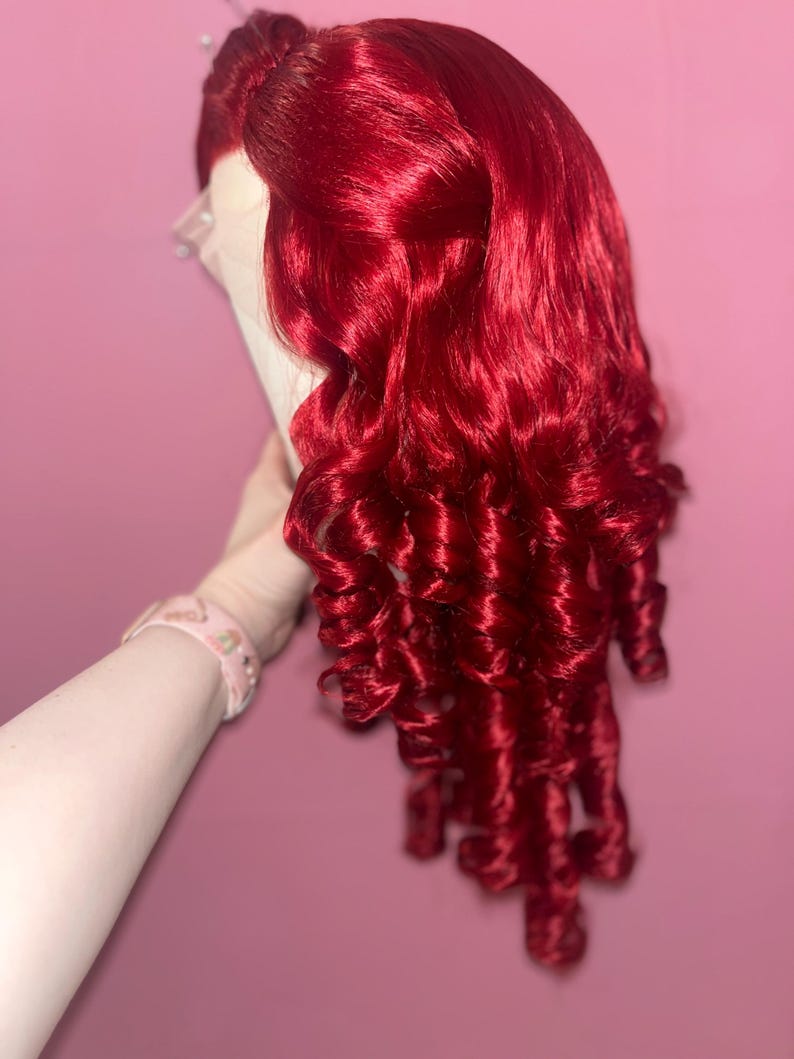 May include: A vibrant, deep red wig featuring long, voluminous hair styled with tight, defined ringlet curls. The hair is parted on the side, showcasing a lace front construction at the hairline, with the curls cascading down in a layered, textured fashion.