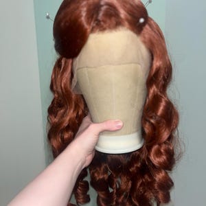 May include: A long, vibrant auburn wig featuring voluminous, loose curls that cascade down the sides. The wig is styled with a prominent, rounded bouffant section at the crown and includes a sheer lace front panel visible at the hairline.