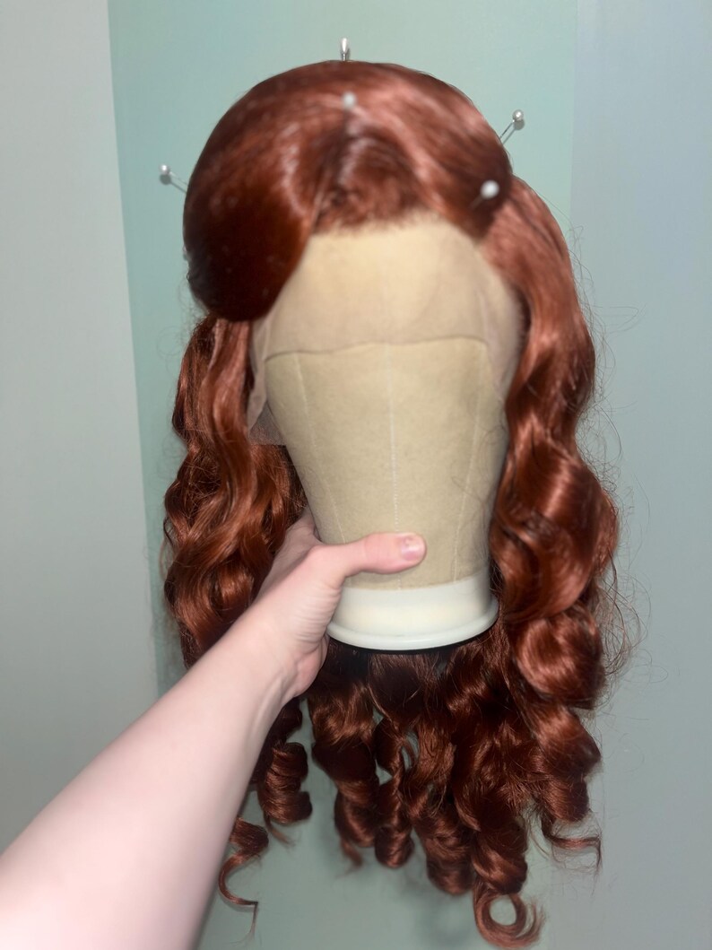 May include: A long, vibrant auburn wig featuring voluminous, loose curls that cascade down the sides. The wig is styled with a prominent, rounded bouffant section at the crown and includes a sheer lace front panel visible at the hairline.