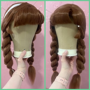 May include: A brown wig styled with two long, thick braids and a fringe. The wig is displayed on a mannequin head, held by a person. The wig has a white headband and is secured with pins. The braids are tied with small brown elastics.