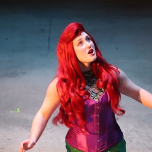 May include: A performer wearing a long, vibrant red wig, a purple corset top with a metallic scale-patterned neckline, and a green skirt. The individual is looking upward with an open mouth, portraying a character in a theatrical costume.