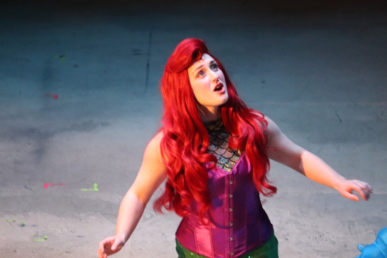 May include: A performer wearing a long, vibrant red wig, a purple corset top with a metallic scale-patterned neckline, and a green skirt. The individual is looking upward with an open mouth, portraying a character in a theatrical costume.