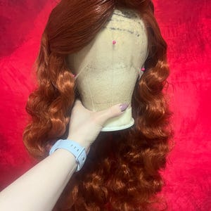 May include: A long, wavy, vibrant copper-red wig styled with a side part and voluminous curls. The wig is secured onto a beige mannequin head with several colourful sewing pins placed along the lace front hairline to hold the hair in place.