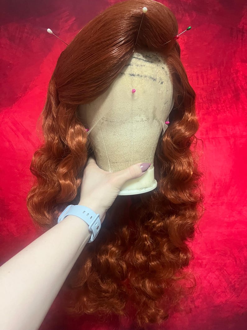 May include: A long, wavy, vibrant copper-red wig styled with a side part and voluminous curls. The wig is secured onto a beige mannequin head with several colourful sewing pins placed along the lace front hairline to hold the hair in place.