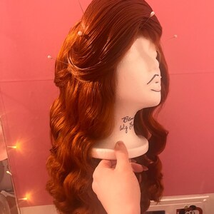 May include: A long, wavy, vibrant copper-red wig styled with a voluminous crown and secured with several white-tipped sewing pins. The wig is displayed on a white foam mannequin head featuring the handwritten text Blow Wig Bar on the neck area.