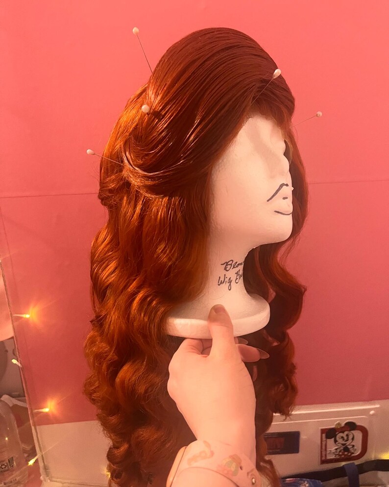 May include: A long, wavy, vibrant copper-red wig styled with a voluminous crown and secured with several white-tipped sewing pins. The wig is displayed on a white foam mannequin head featuring the handwritten text Blow Wig Bar on the neck area.