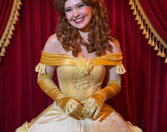 May include: A yellow off-the-shoulder ball gown with a fitted bodice featuring gold glitter scrollwork and a full skirt with rose accents. The ensemble includes matching elbow-length velvet gloves and a hairstyle with a high bun and loose curls.