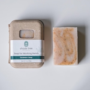 May include: A bar of soap in a cardboard box. The soap is light pink with brown flecks. The box is brown with a green label that says "shade tree naturals" and "Soap for Working Hands Gardener's Soap Net Wt. 5 oz (148g)"