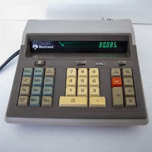May include: A vintage Rockwell 330 calculator with a gray and beige color scheme. The calculator displays the number 80,085 in green digits. It features a variety of function keys and a red minus key.