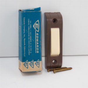 May include: A brown doorbell button with a white push button and two screws. The box it came in is blue with the text "E EDWARDS THIS SYMBOL IS YOUR GUARANTEE OF THE BEST IN SIGNALING, COMMUNICATION AND PROTECTION EQUIPMENT Edwards Company, Inc., Norwalk, Connecticut"
