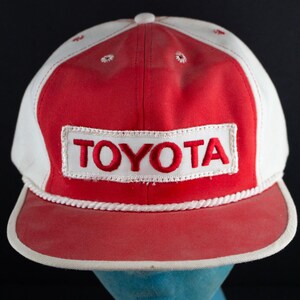 May include: A vintage baseball cap with a red crown and brim, and a white top. The word "TOYOTA" is embroidered in red on a white rectangular patch on the front. The cap has a white rope detail along the brim.