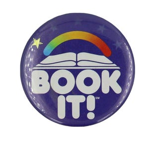 May include: A circular purple pinback button featuring a white open book graphic topped by a colorful rainbow arch. The text BOOK IT! is printed in large white capital letters below the book, with small stars surrounding the design.