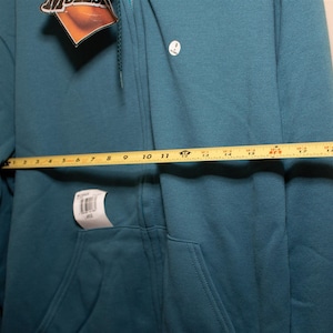 May include: A teal blue zip-up hoodie with a small white logo on the left chest. The hoodie is laid flat and measured with a yellow tape measure, showing a measurement of 17 inches.