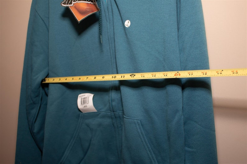 May include: A teal blue zip-up hoodie with a small white logo on the left chest. The hoodie is laid flat and measured with a yellow tape measure, showing a measurement of 17 inches.