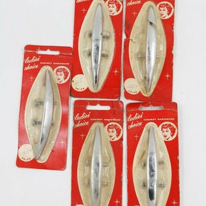 May include: A collection of vintage cabinet hardware pulls on red and cream-colored packaging. The pulls are silver-toned and oval-shaped. The packaging includes the text "Ladies' Choice CABINET HARDWARE".