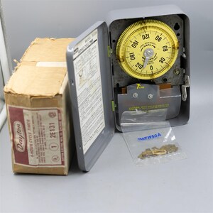 May include: A Dayton 4-hour cycle timer in a gray metal enclosure with an open door, featuring a yellow circular dial marked 0 to 210 minutes. The unit includes a cardboard box with product labels and a small plastic bag containing metal parts.