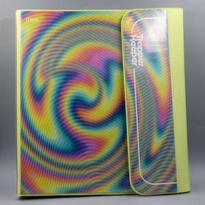 May include: A Trapper Keeper binder with a swirling, colorful design in shades of yellow, pink, blue, and green. The binder is open, revealing the interior. The text "Trapper Keeper" is visible on the spine.