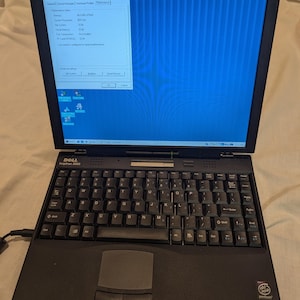 May include: A black Dell Inspiron 3000 laptop with a QWERTY keyboard, a trackpad with two buttons, and a screen displaying the Windows System Properties window. The laptop features an Intel Pentium processor sticker and a Windows 95 logo sticker.