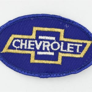 May include: An oval-shaped embroidered patch featuring a blue background with a gold-colored Chevrolet bowtie logo outline. The word CHEVROLET is stitched in white capital letters across the center of the gold bowtie emblem on the blue fabric.