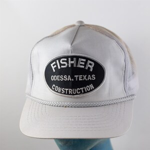 May include: A light gray trucker hat featuring a mesh back and a braided cord across the base of the crown. An oval black patch is centered on the front with white embroidered text that reads FISHER ODESSA, TEXAS CONSTRUCTION.