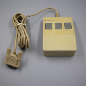 May include: A vintage beige computer mouse with a rectangular body and three grey rectangular buttons. The device features the word LOGITECH printed on the top surface and includes a long beige cord ending in a multi-pin serial connector plug.