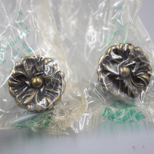 May include: Two antique gold-toned cabinet knobs with a floral design. The knobs are packaged in clear plastic bags.