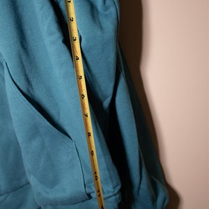 May include: A teal blue hoodie with a pocket on the front. A measuring tape is placed along the sleeve, showing a measurement of 11 inches.