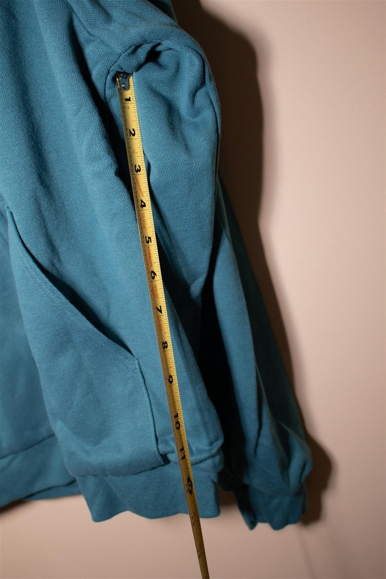 May include: A teal blue hoodie with a pocket on the front. A measuring tape is placed along the sleeve, showing a measurement of 11 inches.