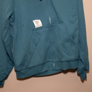 May include: A teal blue zip-up hoodie with a pocket on the front. The hoodie has a tag attached to it with a barcode and the word "Large" printed on it.