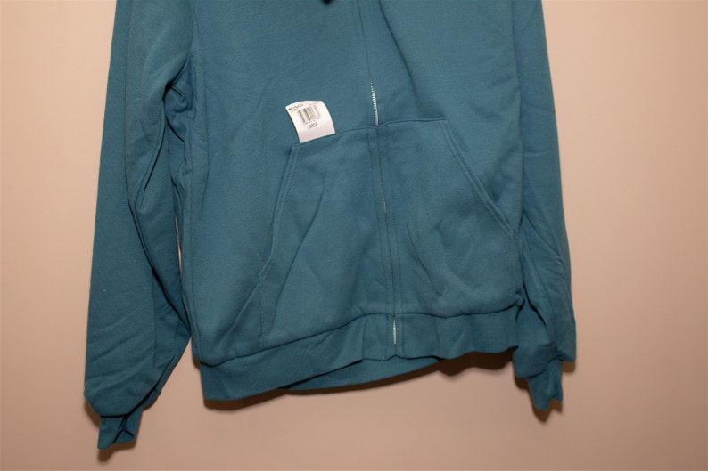 May include: A teal blue zip-up hoodie with a pocket on the front. The hoodie has a tag attached to it with a barcode and the word "Large" printed on it.