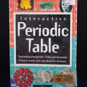 May include: A rectangular CD-ROM box titled Interactive Periodic Table with a background of a periodic table grid in red, white, and blue. The cover features scientific imagery, a portrait of Marie Curie, a molecular model, and text descriptions.