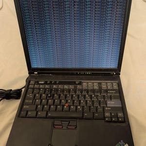 May include: A black IBM ThinkPad laptop with a QWERTY keyboard and a red pointing stick in the center of the keys. The screen displays a vertical pattern of blue and black digital artifacts, indicating a display or graphics processing error.