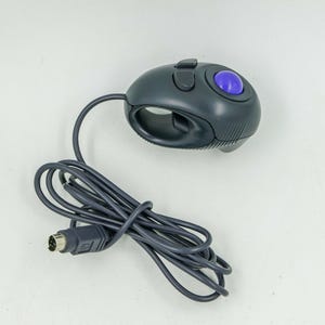 May include: A dark gray handheld trackball mouse featuring a prominent purple ball, a top-mounted button, and a trigger-style button on the side. The device is connected to a long, coiled dark gray cable that terminates in a round multi-pin connector.