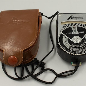 May include: An Adams light meter with a silver and black face featuring dials for film ASA speed and shutter speed, connected by a black cord to a brown leather carrying case with a snap closure and an embossed Adams logo on the front flap.