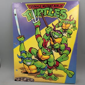 May include: A rectangular Teenage Mutant Ninja Turtles three-ring binder featuring a colorful graphic of the four turtles wielding weapons including a katana, sai, bo staff, and nunchucks. The background is split between purple and yellow diagonal sections.