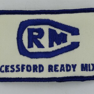 May include: A white patch with blue stitching around the edges. The patch has a blue oval with the letters "RM" inside. Below the oval is the text "CESSFORD READY MIX".