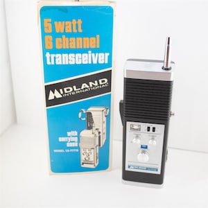 May include: A black and silver Midland International 5 watt 6 channel transceiver with a vertical antenna, control knobs, and a silver faceplate. Beside it stands the original blue and white cardboard box labeled 5 watt 6 channel transceiver.