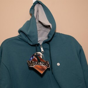 May include: A teal blue zip-up hoodie with a gray fleece lining. The hoodie has a small white tag with the text "Muleskins" on it.