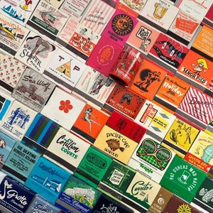 May include: A collection of vintage matchbooks with various designs and text, including restaurant and business advertisements. The matchbooks feature a range of colors and graphics, creating a nostalgic aesthetic. A can of "Tomatoes" is in the center.