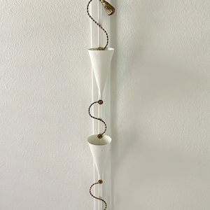 May include: White metal wall planter with three cone-shaped pots hanging from a gold chain with tassels.