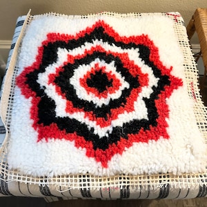 May include: A close-up of a red, black, and white rug with a circular pattern. The rug is on a white canvas with a grid pattern.