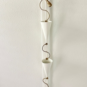 May include: White metal hanging planter with three cone-shaped pots connected by a gold rope with tassels.